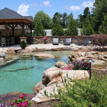 Backyard pool with turquoise water, rock edging, flowers, lounge chairs, and a gazebo under a blue sky