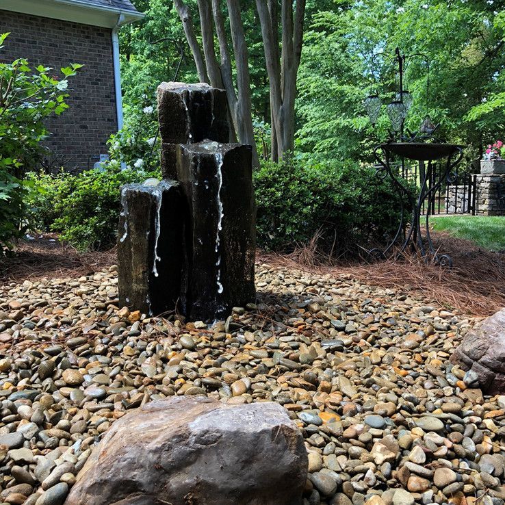 Small tiered black rock fountain trickling water in a pebble garden with greenery and trees.