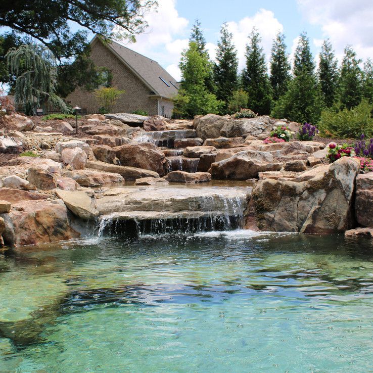 Rock waterfall flowing into a clear turquoise pond with landscaped boulders and trees nearby