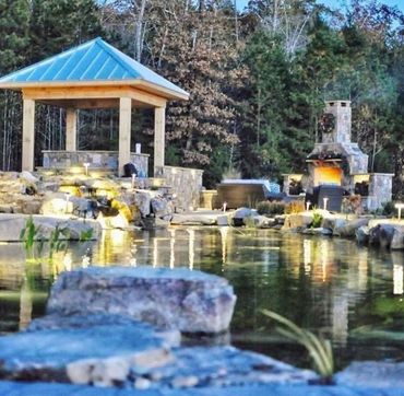 Stone-lined pond with a blue-roof pavilion and glowing outdoor fireplace surrounded by trees