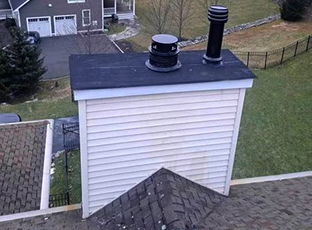 Chimney on a roof with two black flue pipes. The chimney has white siding and a black roof.