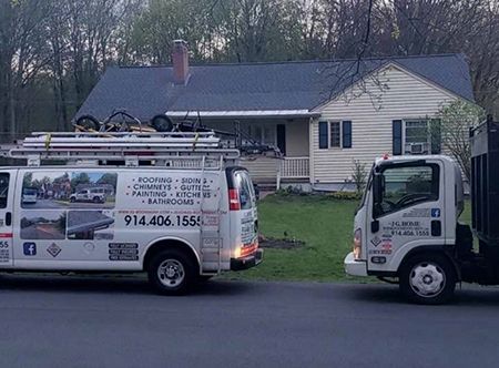 A white work van and truck parked near a house with a dark roof.