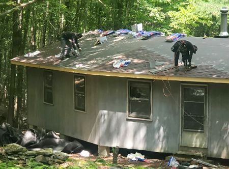 Two people working on a roof of a round house in a wooded area. They appear to be installing new shingles.