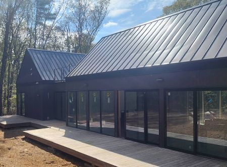 Modern dark-brown house with a metal roof and large glass windows surrounded by trees.