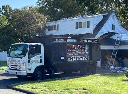 Dump truck parked in front of a house under roof repair. Green grass and trees surround.