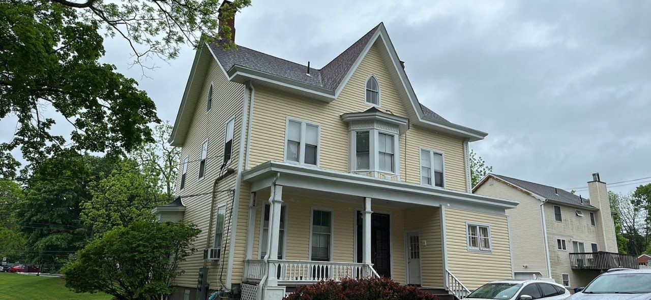 A large yellow house with a porch and cars parked in front of it on a cloudy day.