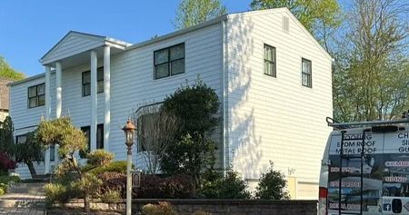 White two-story house with columns and dark-framed windows, surrounded by landscaping and a van on the right.