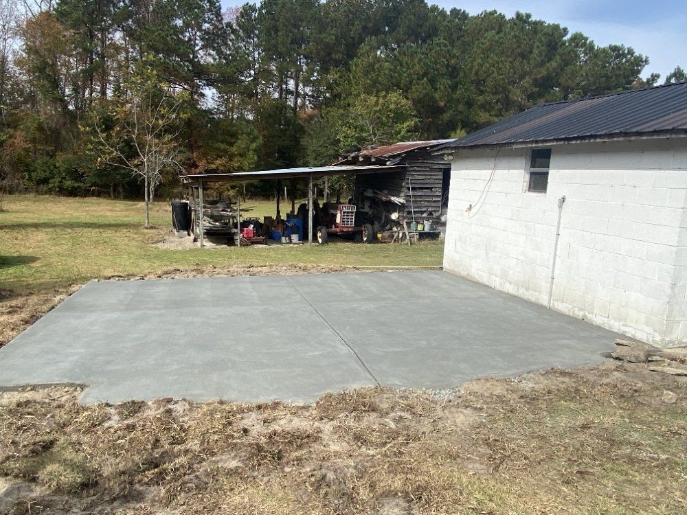 Newly poured concrete patio in a yard next to a weathered building and shed; brown grass and trees in background.