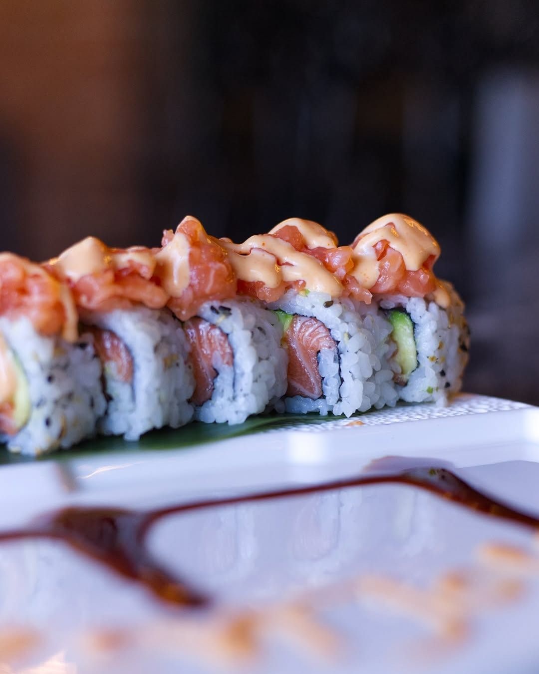 Primo piano di un rotolo di sushi guarnito con salmone piccante e salsa cremosa, adagiato su un piatto irrorato con glassa di soia scura.