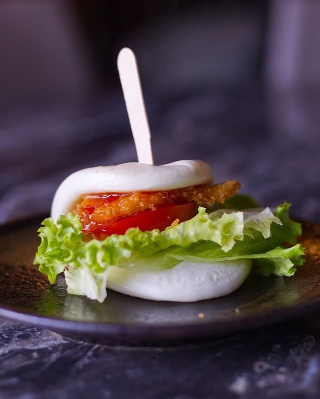 Un panino bao ripieno di una cotoletta di pollo croccante, lattuga fresca e una fetta di pomodoro, tenuto insieme da uno stuzzicadenti di legno.