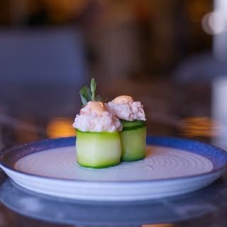 Due antipasti avvolti in cetriolo, guarniti con una crema spalmabile e una piccola decorazione, serviti su un piatto bianco con il bordo blu.