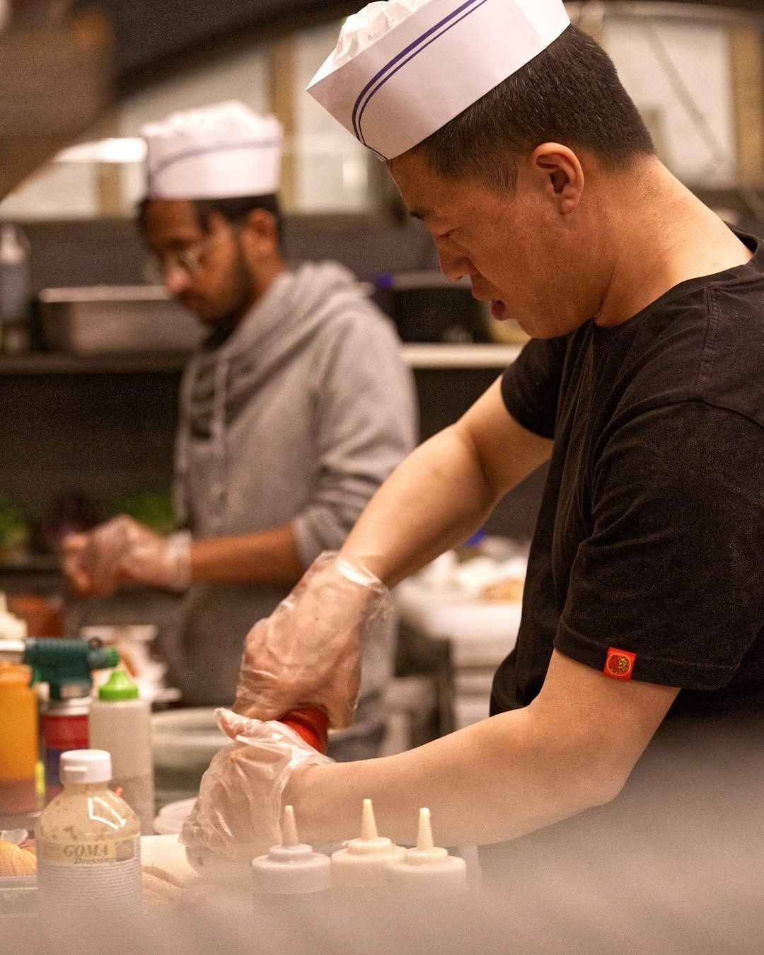 In una cucina di ristorante, due chef con cappelli di carta bianchi e guanti di plastica trasparente lavorano, uno dei quali sta versando la salsa su un piatto.