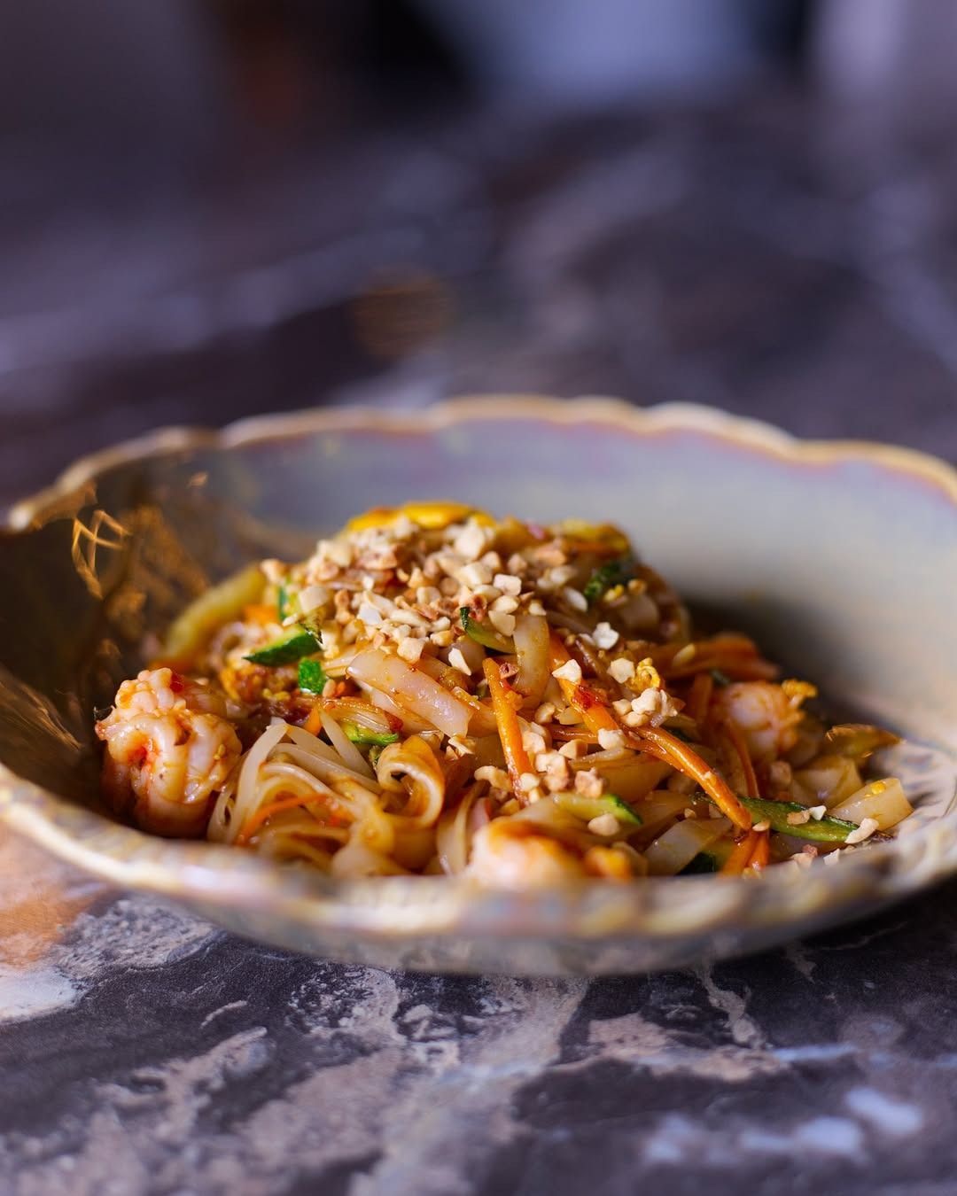 Una ciotola di ceramica poco profonda riempita di Pad Thai, guarnita con arachidi tritate, servita su un piano di lavoro in pietra scura e screziata.