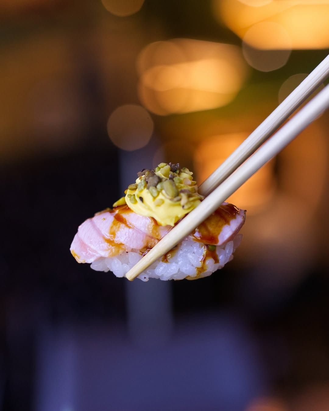 Delle bacchette sorreggono un pezzo di nigiri sushi condito con salsa gialla ed erbe tritate, sullo sfondo di un'immagine sfocata e dorata.