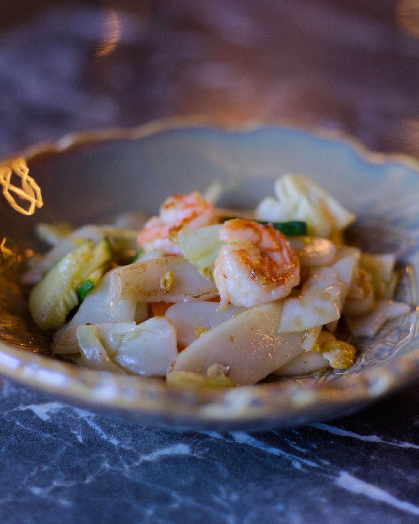 Primo piano di gamberi saltati in padella con cavolo e tagliatelle di riso piatte in una ciotola smerlata su una superficie di marmo scuro.