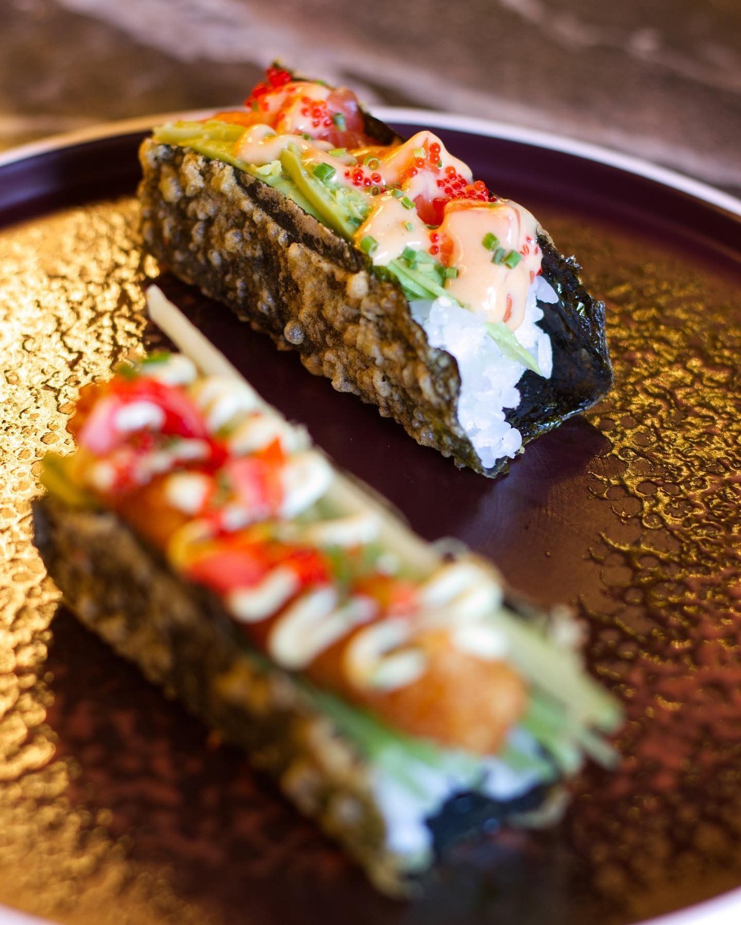 Due tacos di sushi con gusci di nori fritti in tempura, guarniti con frutti di mare, salsa cremosa e decorazioni, serviti su un piatto scuro.