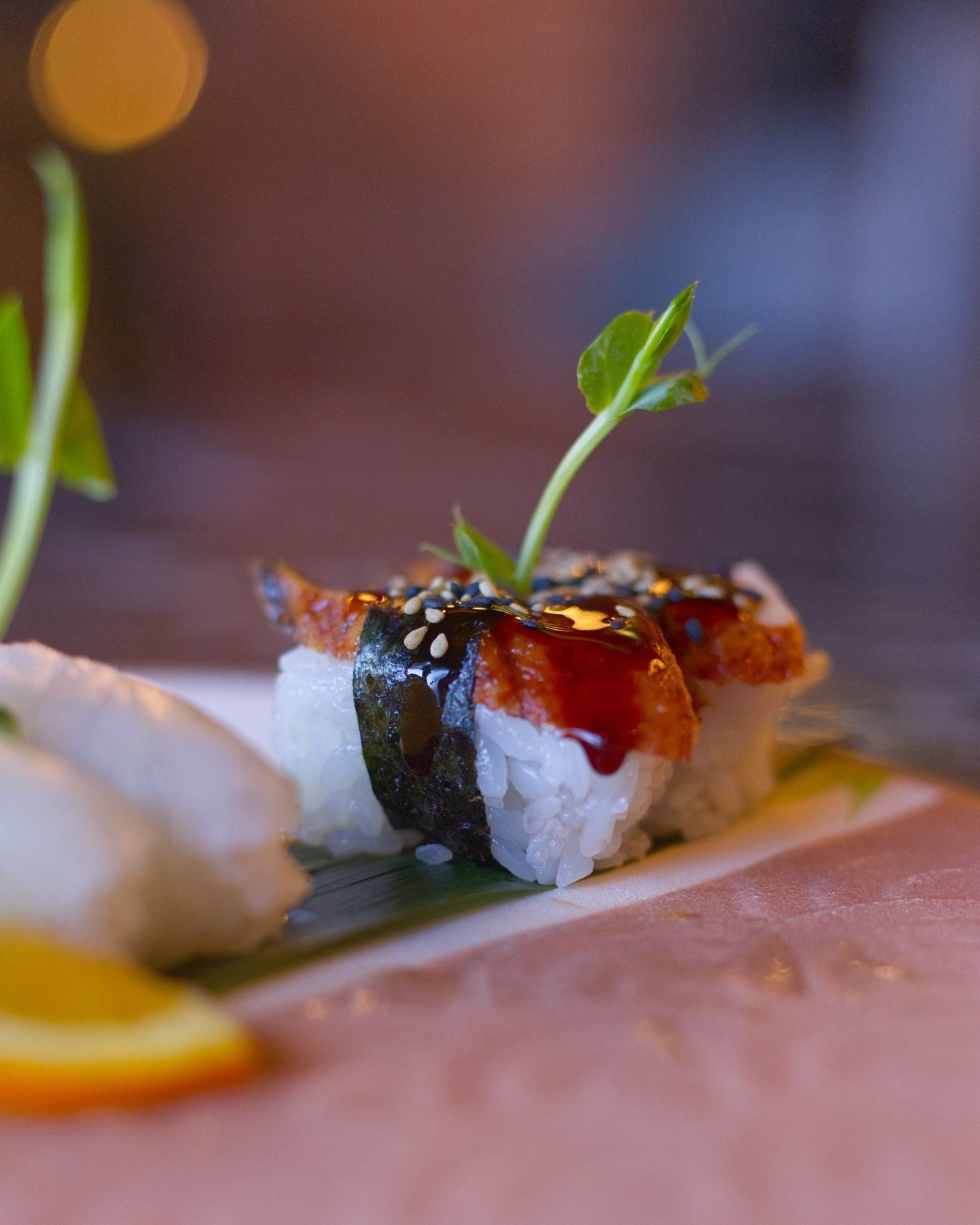 Due pezzi di nigiri di anguilla guarniti con semi di sesamo e un rametto di verde, serviti su una foglia.