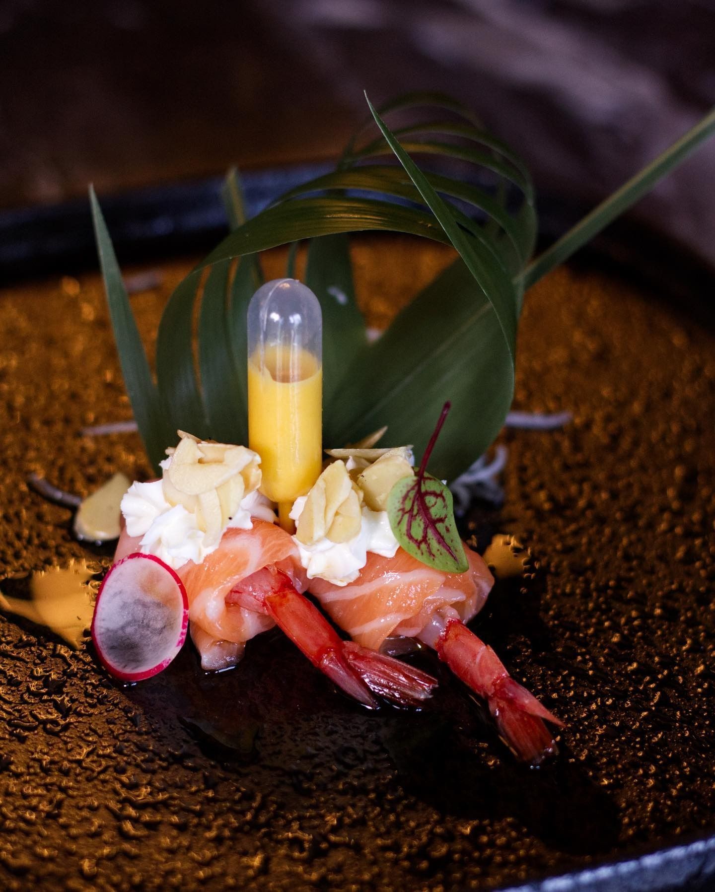 Due gamberi avvolti nel salmone, guarniti con panna bianca, un tocco decorativo e una piccola goccia gialla su un piatto scuro dalla superficie ruvida.