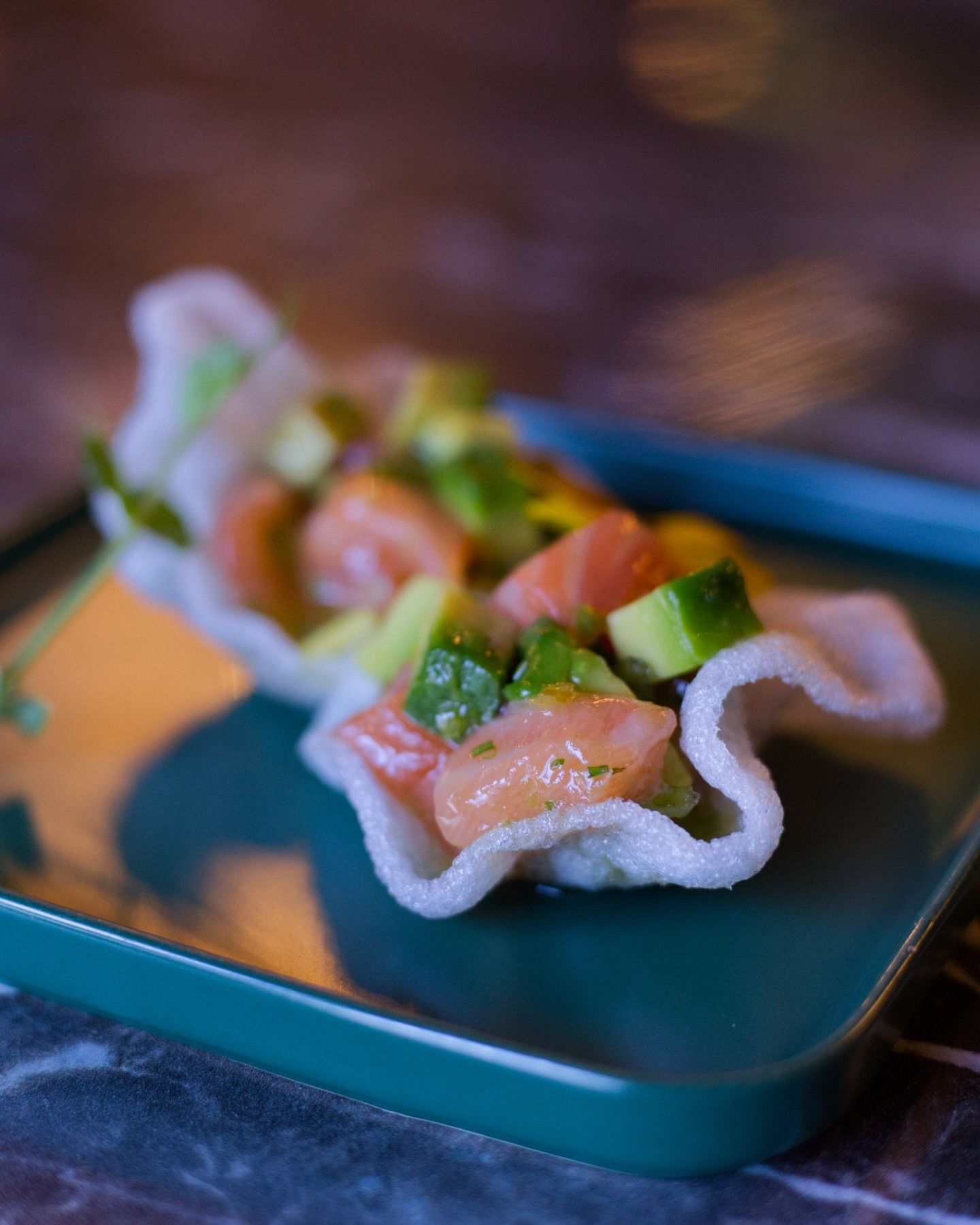 Un piccolo cracker croccante di gamberi ripieno di tartare di salmone e avocado, servito su un piatto quadrato color verde acqua.