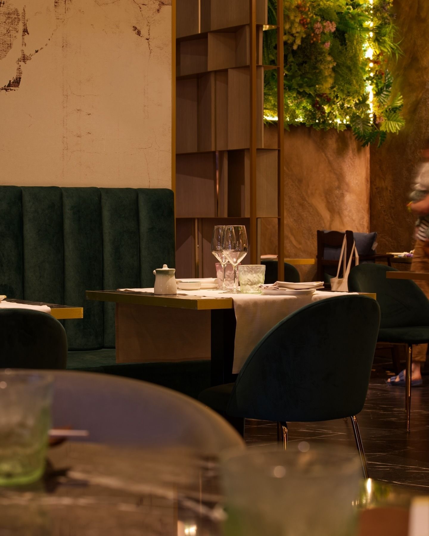 L'interno di un ristorante, illuminato da una luce calda, presenta sedie imbottite di colore verde scuro, un tavolo con bicchieri e decorazioni con piante verdi.