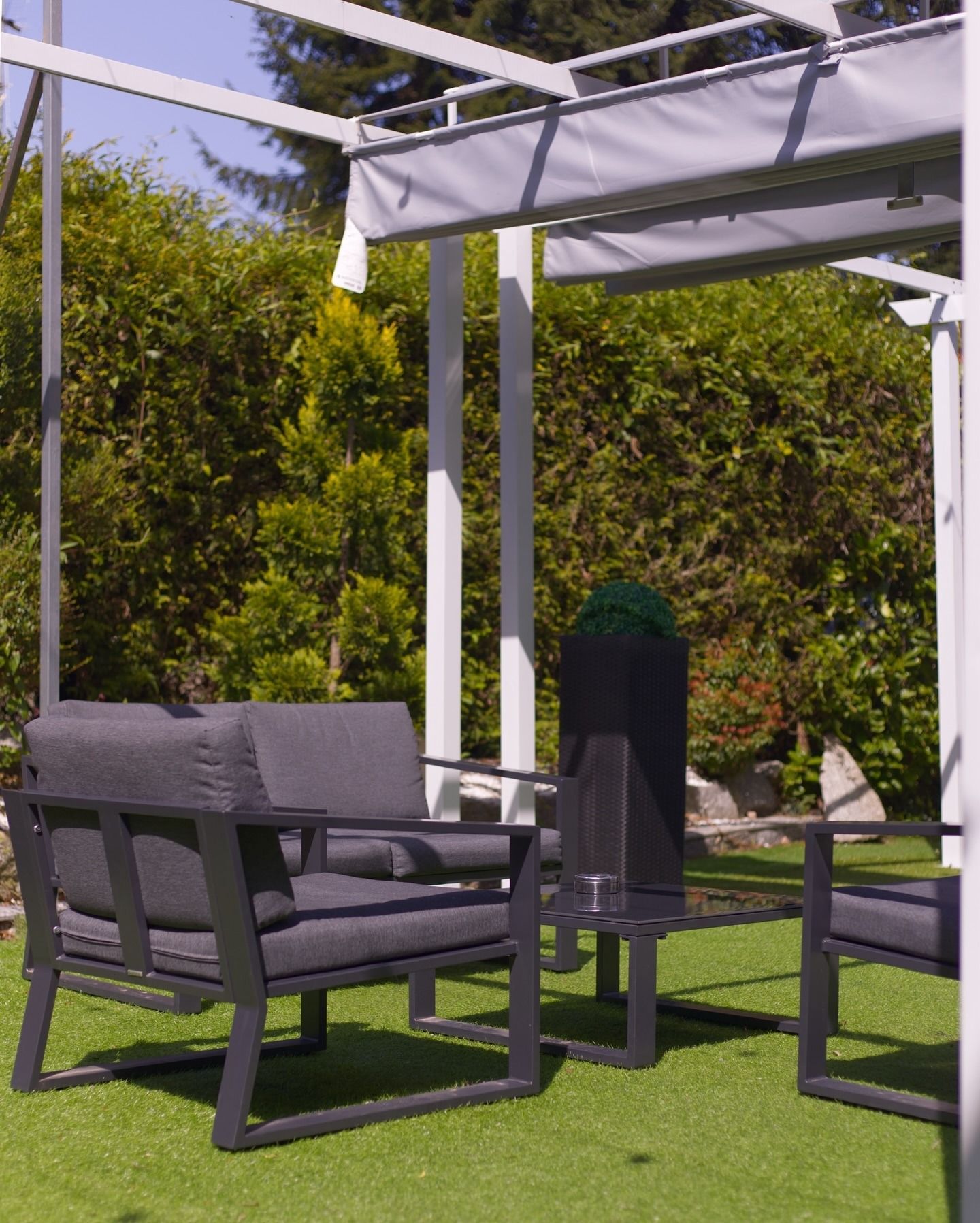 Area patio esterna con sedute imbottite grigio scuro e un tavolo abbinato sotto un pergolato bianco su erba artificiale.
