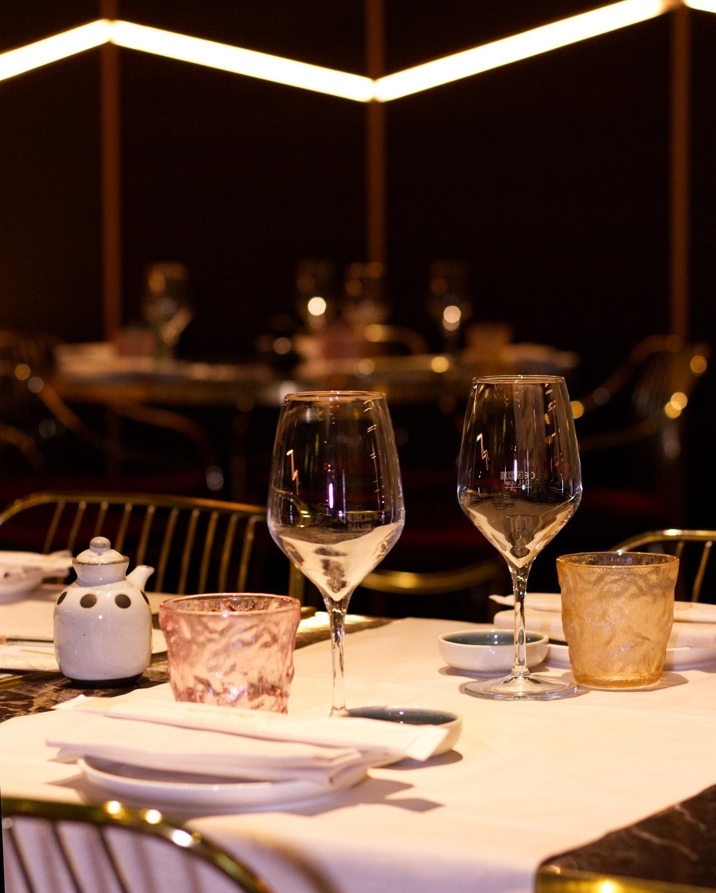 Due calici da vino, bicchieri dalla superficie ruvida, e un barattolo di ceramica per condimenti su un tavolo da ristorante apparecchiato con una tovaglia bianca.