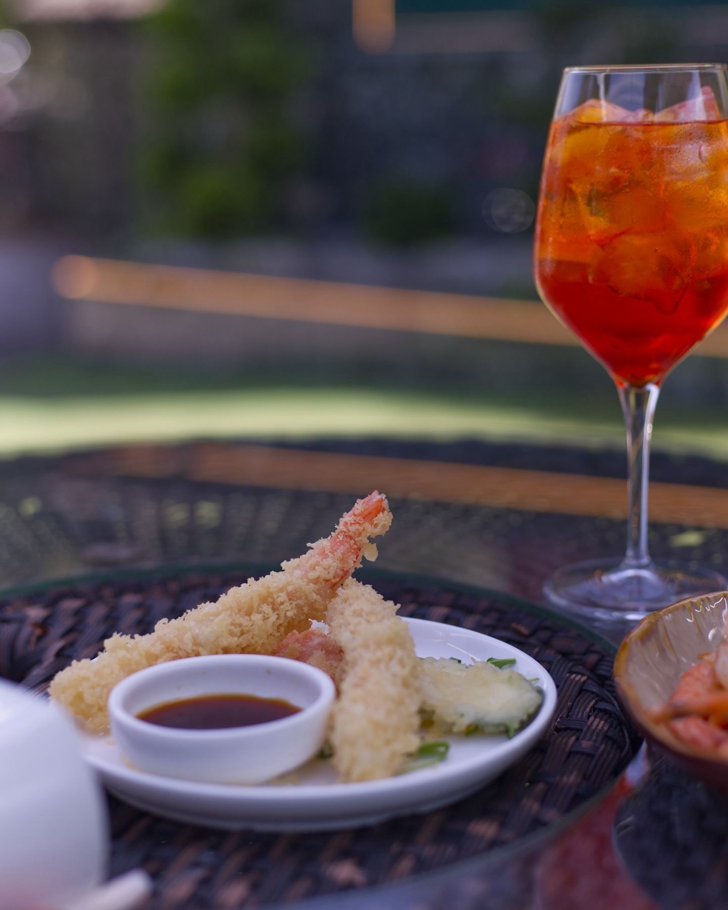 Un piatto di tempura di gamberi con salsa d'accompagnamento, accanto a un bicchiere di cocktail arancione brillante, su un tavolo di vimini all'aperto.
