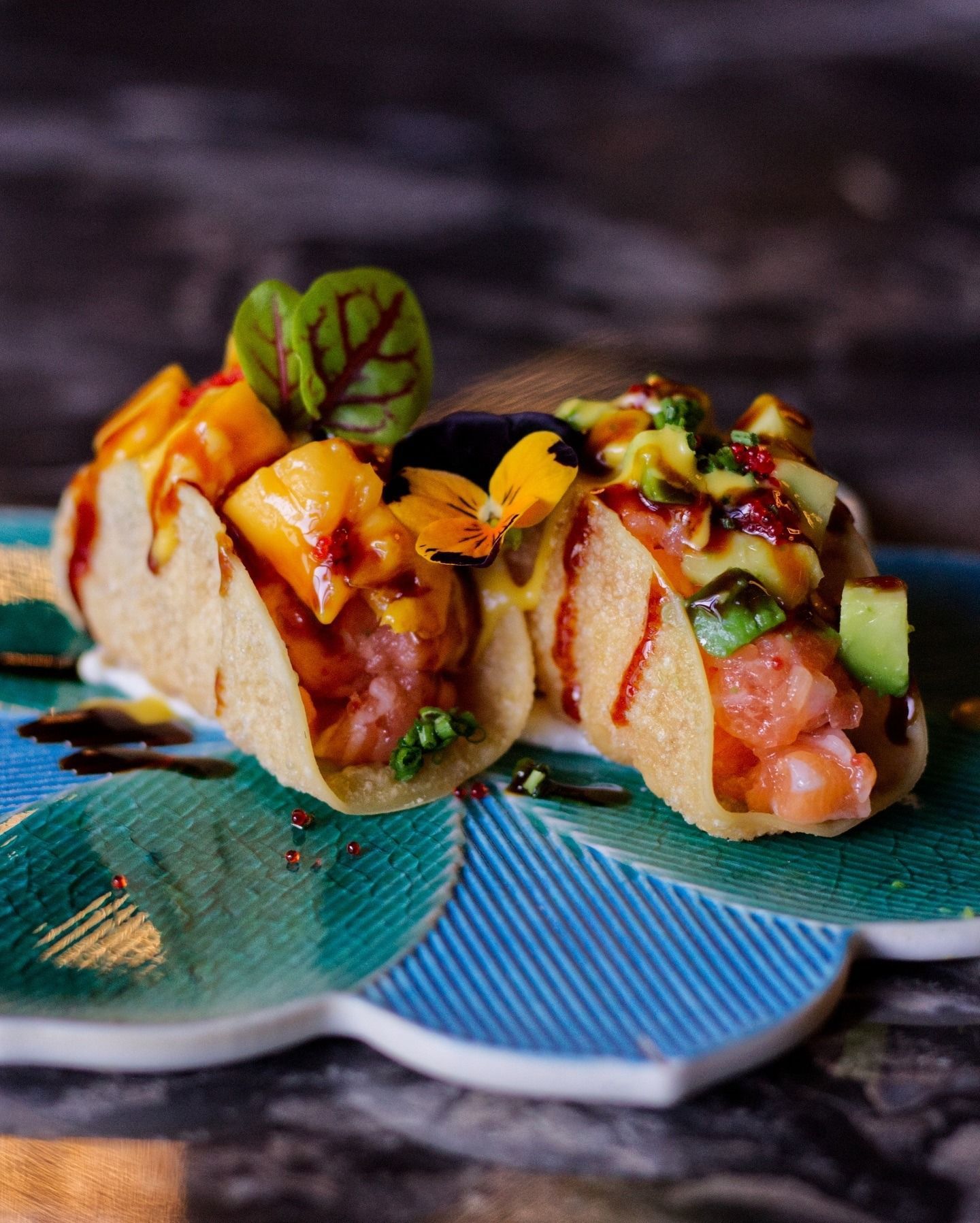 Due tacos di tartare di tonno guarniti con mango, avocado e fiori edibili, serviti su un piatto decorativo con motivi floreali blu e verdi.