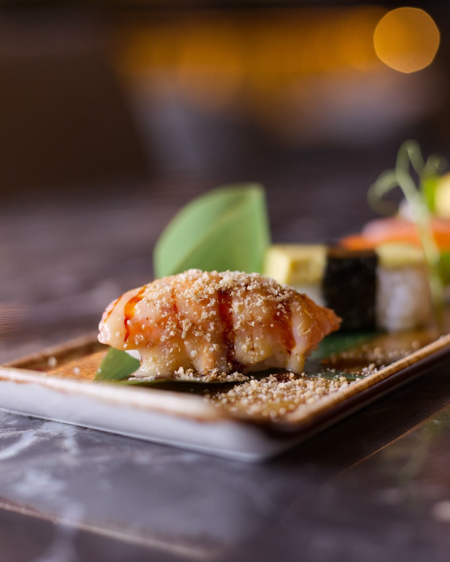 Un piatto di sushi con un pezzo di salmone scottato, condito con salsa e semi di sesamo, servito su una superficie di pietra scura.