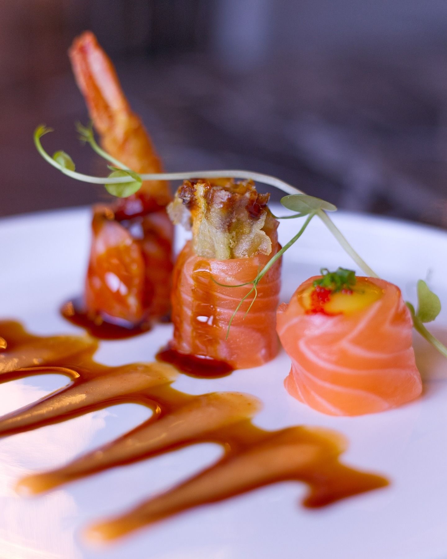 Tre pezzi di sushi avvolti nel salmone, serviti su un piatto bianco con striature artistiche di glassa di soia scura e guarnizione di microverdure.