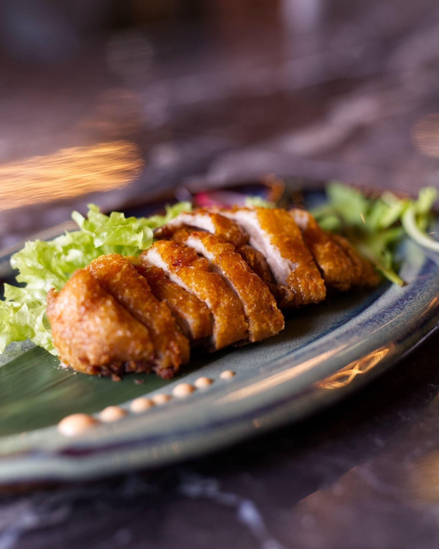 Cotoletta di pollo fritta a fette, servita su una foglia verde e un piatto di ceramica, con contorno di lattuga e piccole gocce di salsa.