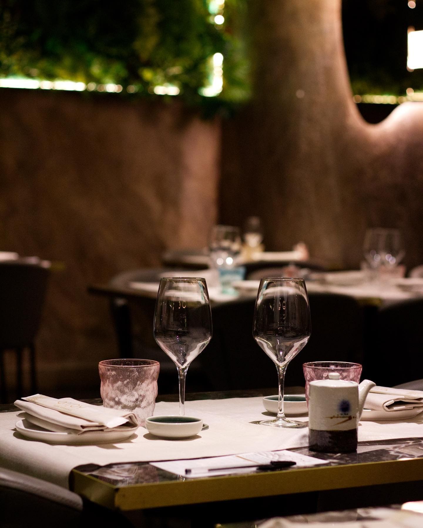 Un ambiente di ristorante elegante e con luci soffuse, con un tavolo apparecchiato per due, completo di bicchieri da vino, tovaglioli e piattini.