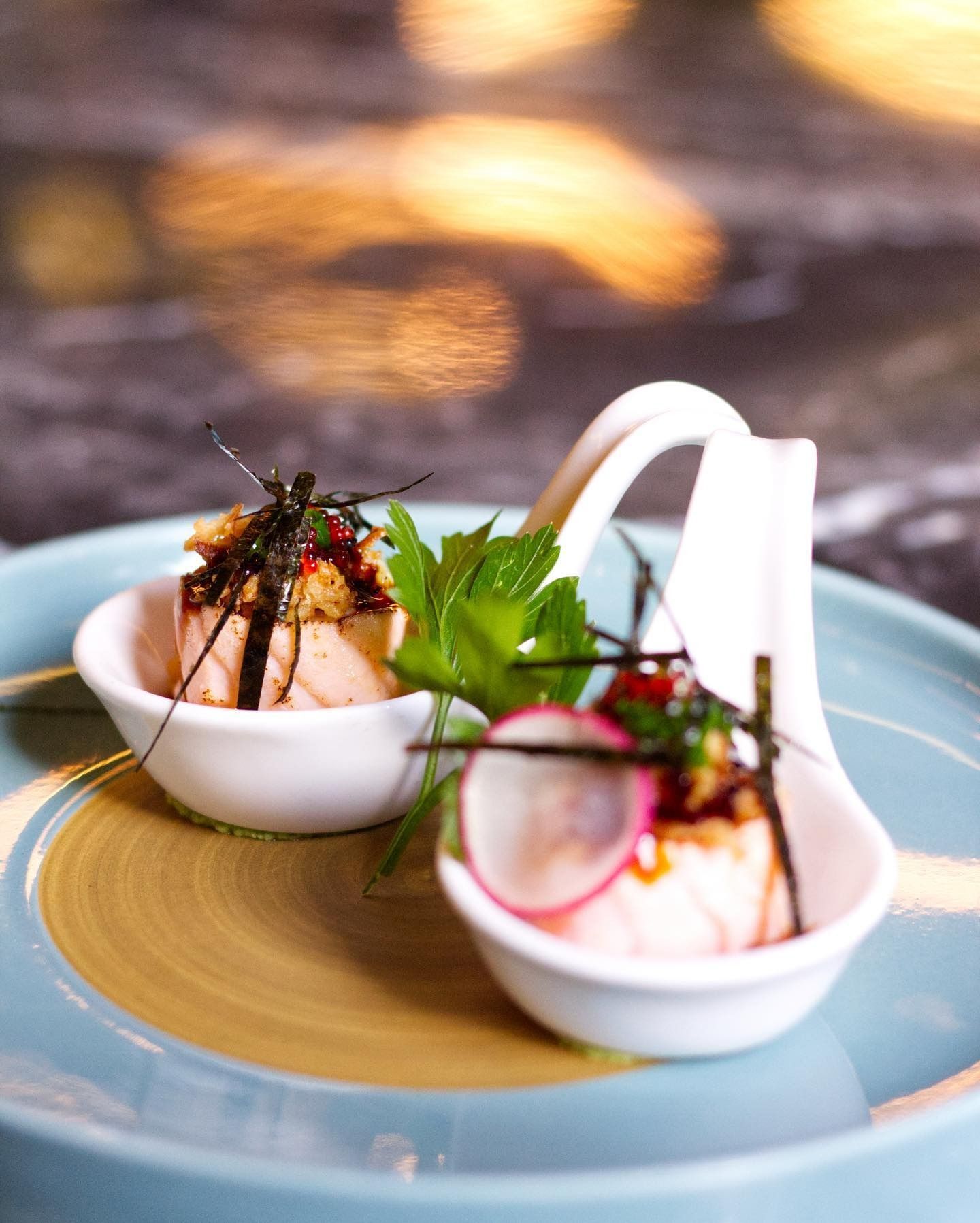 Due porzioni di pesce scottato, guarnite con contorno e fettine di ravanello, servite in cucchiai bianchi su un piatto blu.