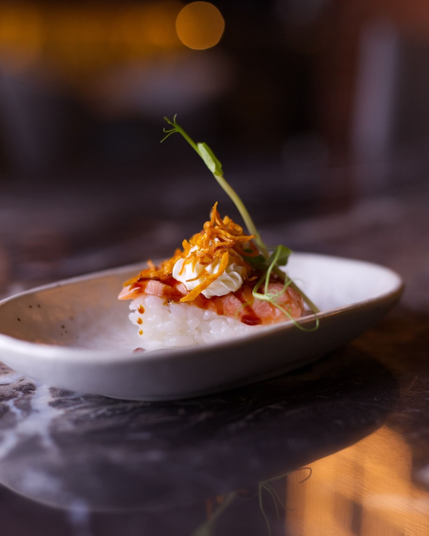 Un singolo pezzo di sushi guarnito con cipolle fritte croccanti, formaggio cremoso e un germoglio di piselli, servito su un piatto di ceramica.