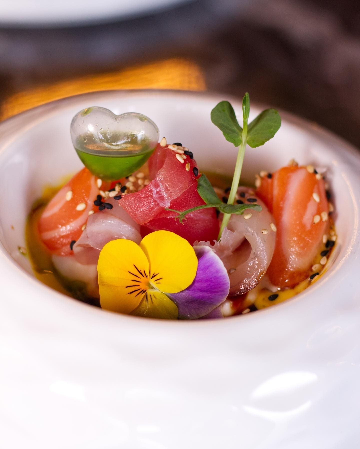 Una ciotola gourmet di sashimi con tonno, salmone, fiori eduli, una goccia di olio alle erbe a forma di cuore e una guarnizione di germogli di piselli.
