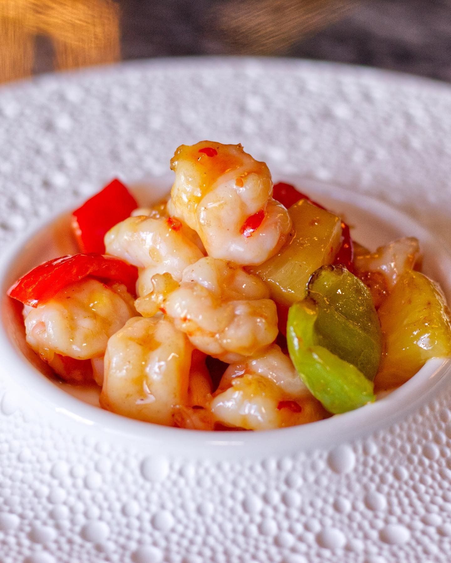 Una piccola ciotola bianca piena di gamberi saltati in padella con peperoni rossi, peperoni verdi e pezzi di ananas glassati.