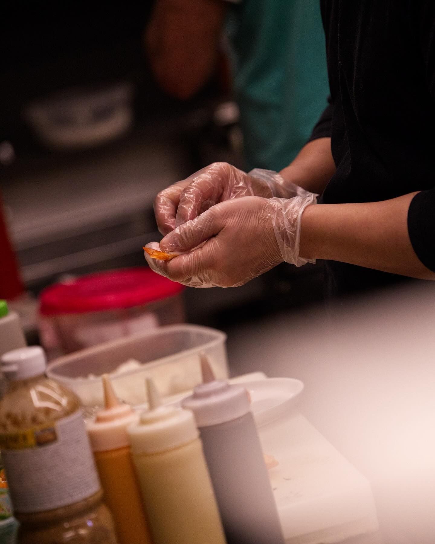Una persona con indosso guanti di plastica trasparente usa le mani per preparare il cibo, con bottiglie di condimenti in primo piano.