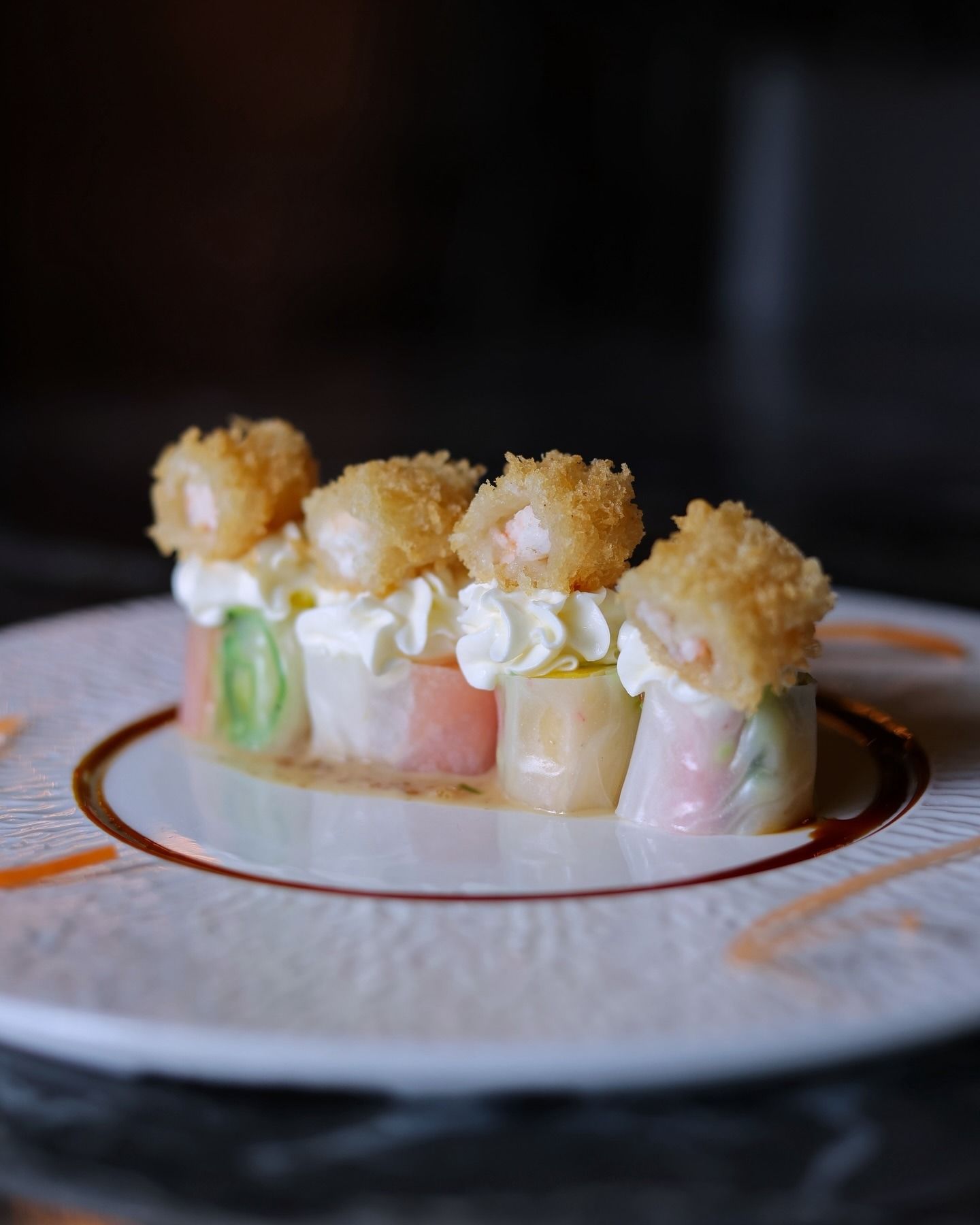Quattro pezzi di sushi guarniti con cucchiaiate di panna e gamberi fritti croccanti, disposti su un piatto bianco con salsa.