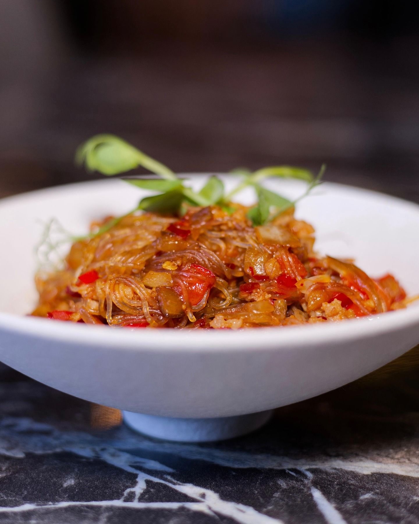 Una ciotola bianca di vermicelli di soia saltati in padella con peperoni rossi e cipolle, guarnita con un rametto di verdure, su un tavolo di marmo.