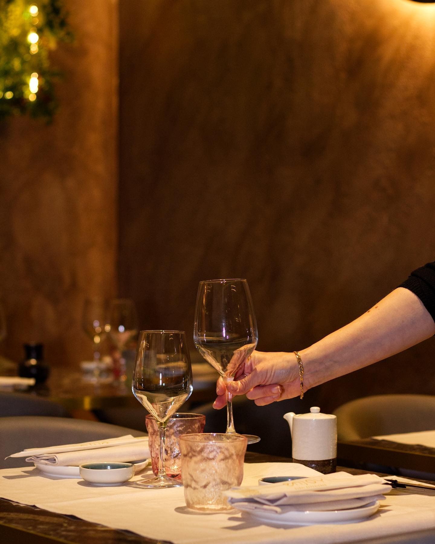 Una mano sistema un calice di vino su un tavolo di ristorante apparecchiato con tovaglie bianche, bicchieri e piccoli piatti di ceramica.