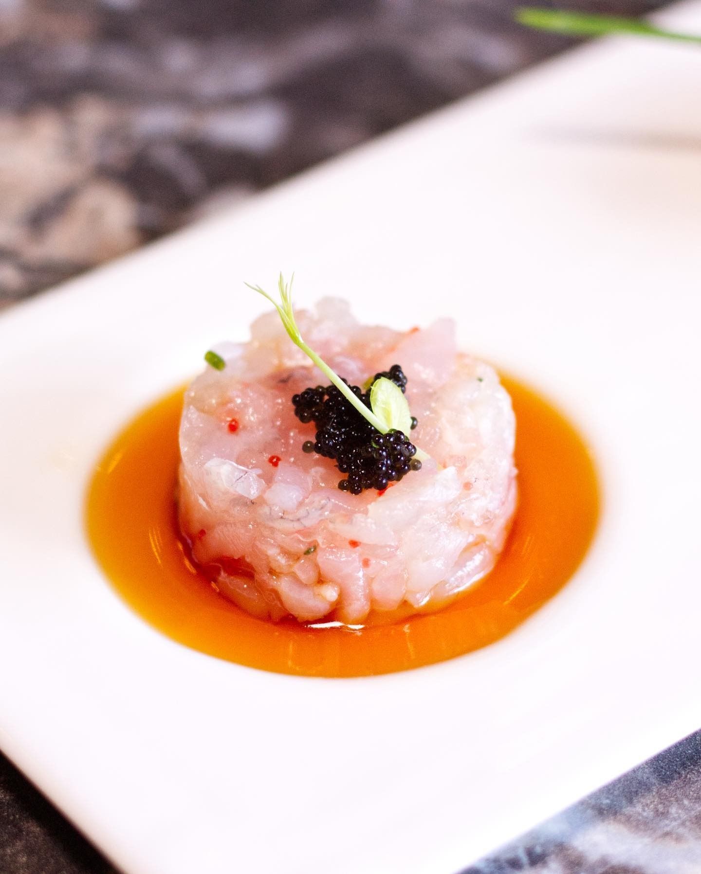 Un mucchietto rotondo di pesce crudo tagliato finemente, guarnito con caviale nero e un germoglio verde, servito in una pozza di salsa arancione.