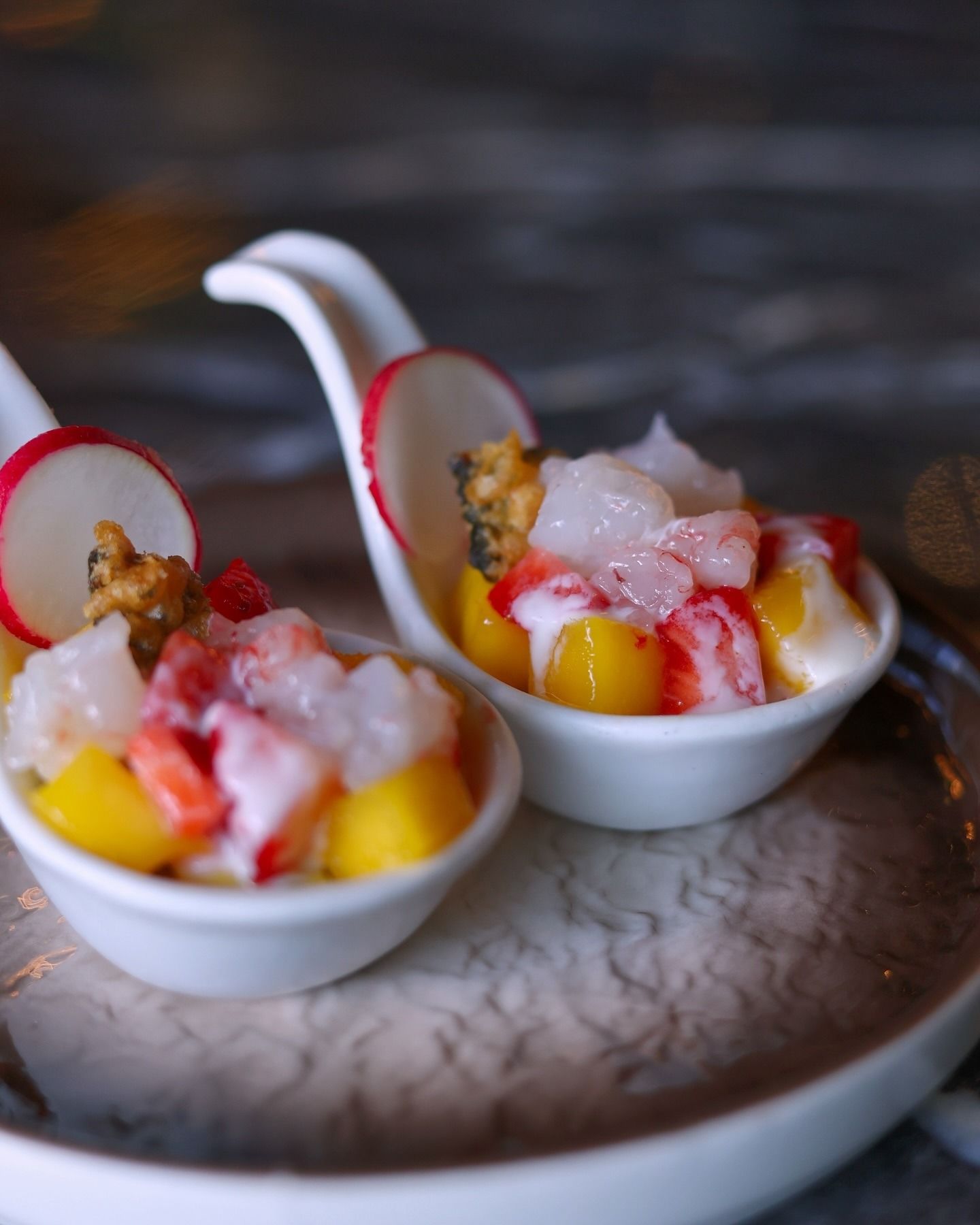 Due cucchiai da antipasto in ceramica che reggono un'insalata colorata di frutti di mare e mango, guarnita con una sottile fetta di ravanello, su un piatto grigio.