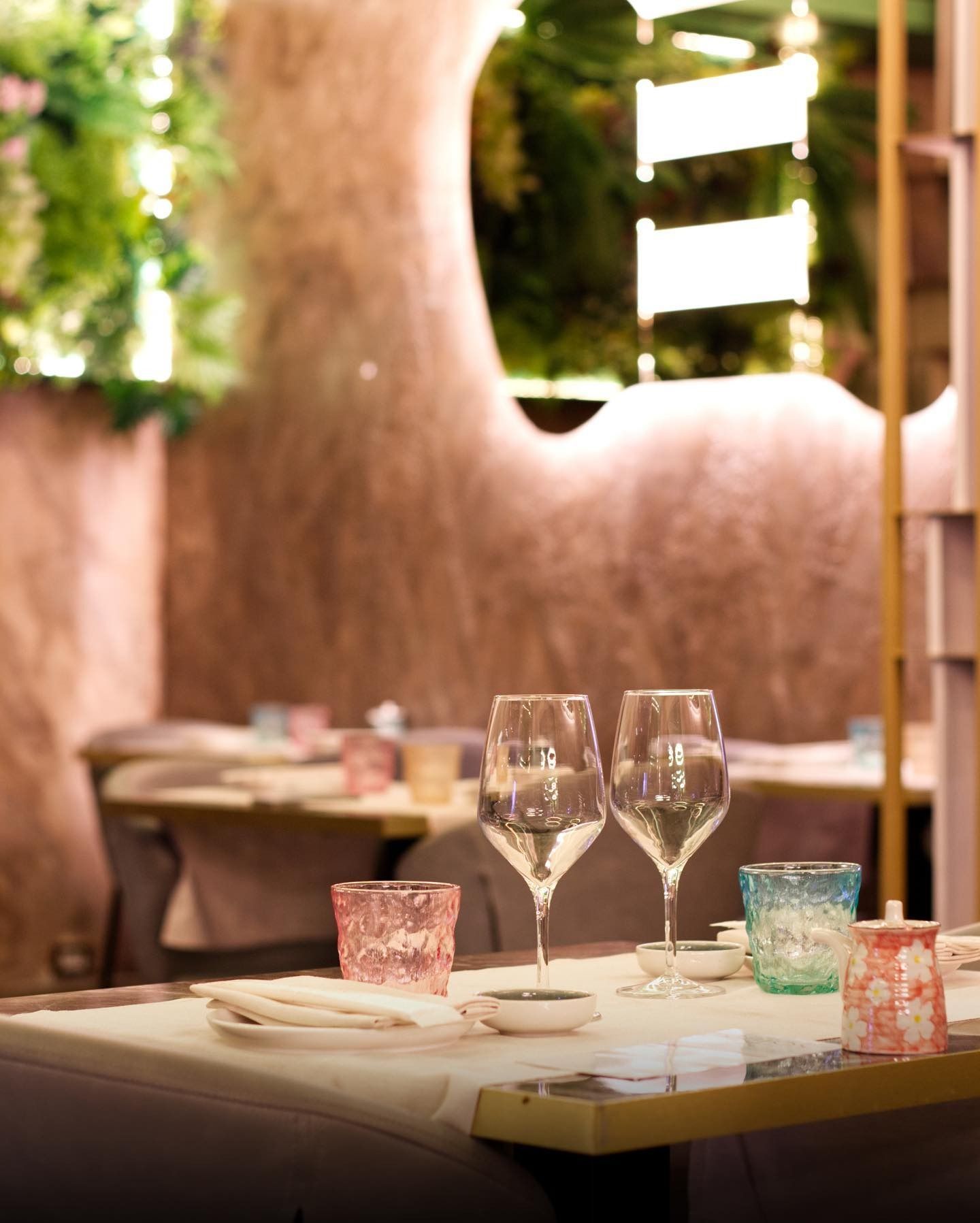 Un tavolo apparecchiato per cenare, con calici da vino e bicchieri decorati, in un ristorante con pareti ruvide di colore rosa-marrone.