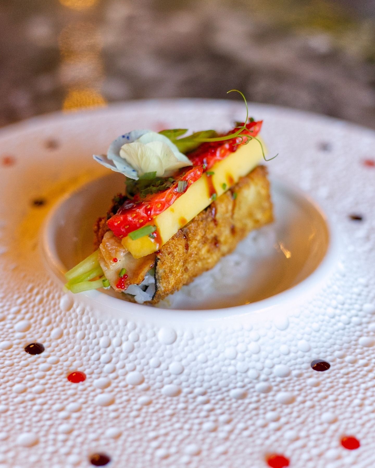 Un pezzo di sushi gourmet guarnito con peperone arrostito e un fiore edibile, servito su un piatto bianco dalla superficie ruvida con puntini.