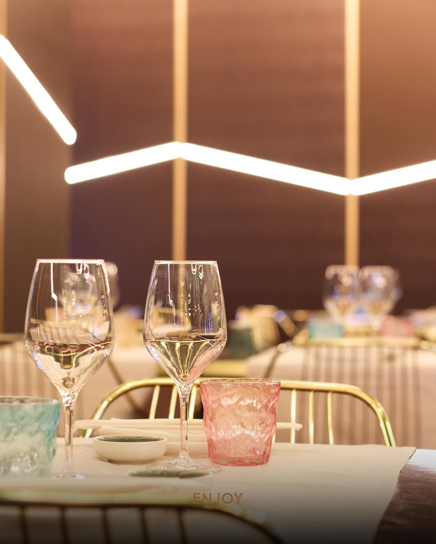 Elegante allestimento da ristorante con calici da vino in cristallo, un bicchiere rosa dalla texture particolare e una morbida illuminazione ambientale moderna.