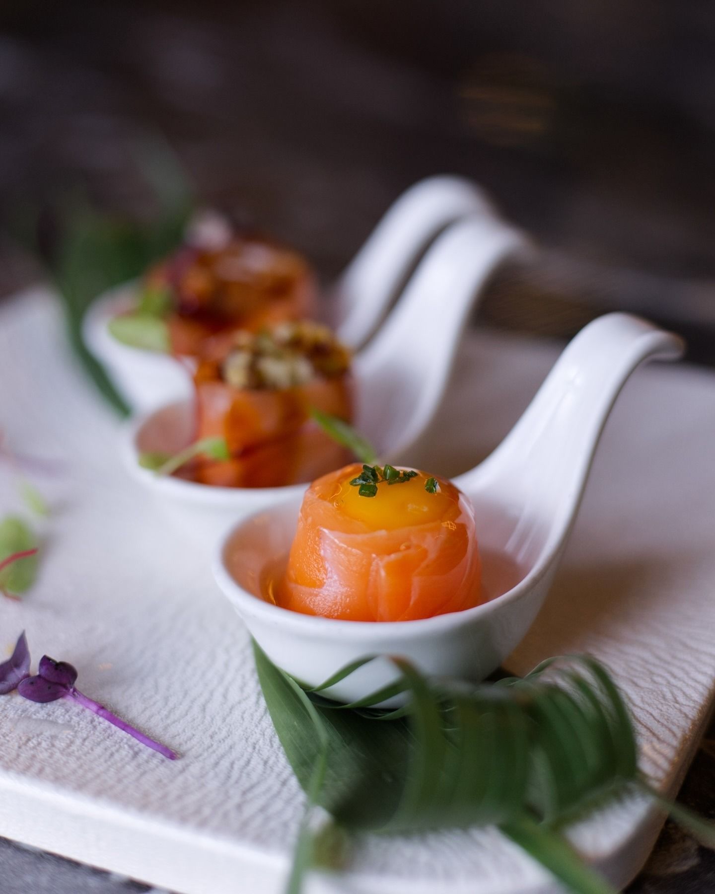 Tre cucchiaini da degustazione bianchi contengono ciascuno un pezzo arrotolato di salmone affumicato sormontato da un tuorlo di un giallo brillante e da erbe aromatiche.