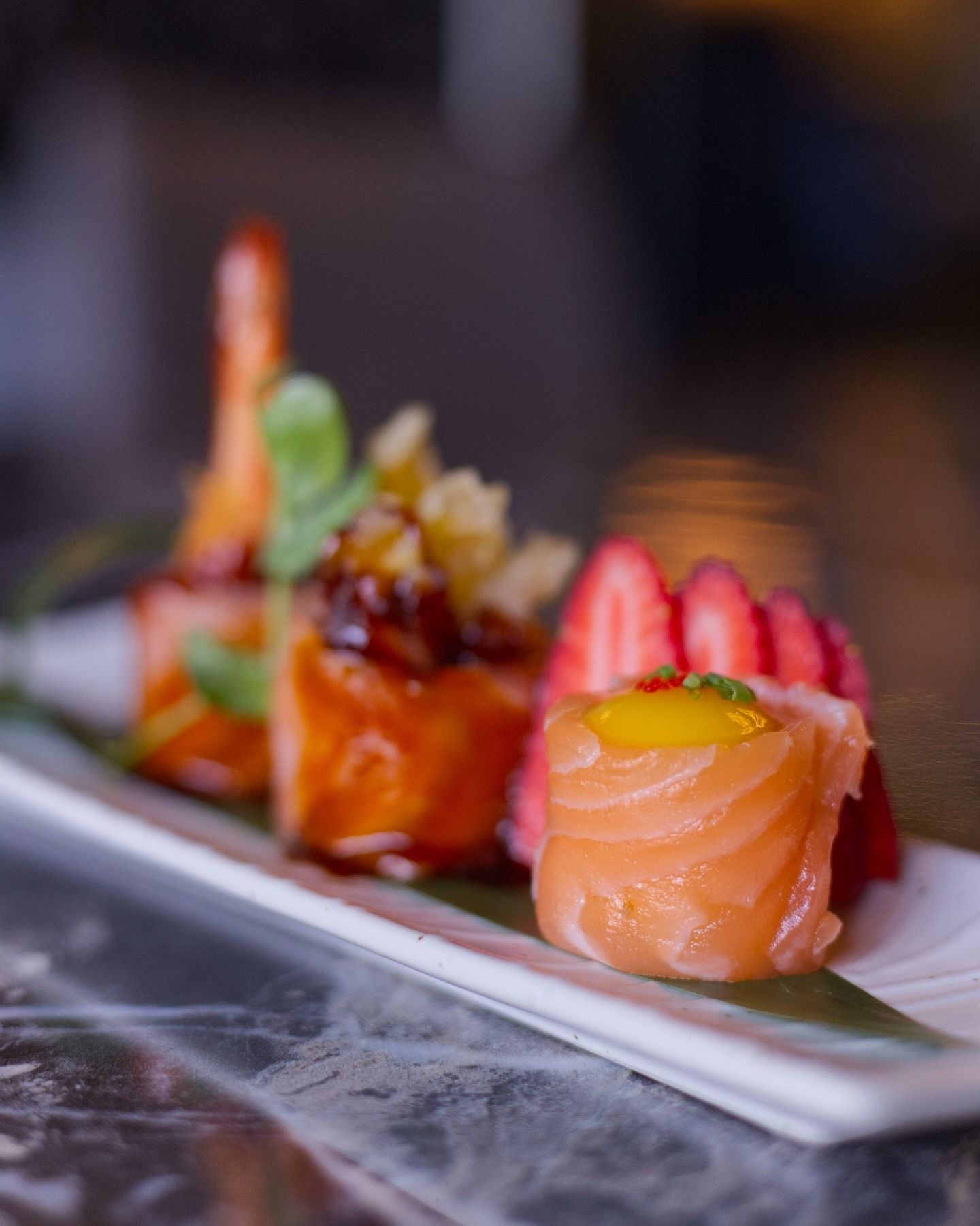 Su un piatto rettangolare bianco sono disposti tre pezzi di sushi, tra cui nigiri di salmone guarniti con mango e fragole fresche.