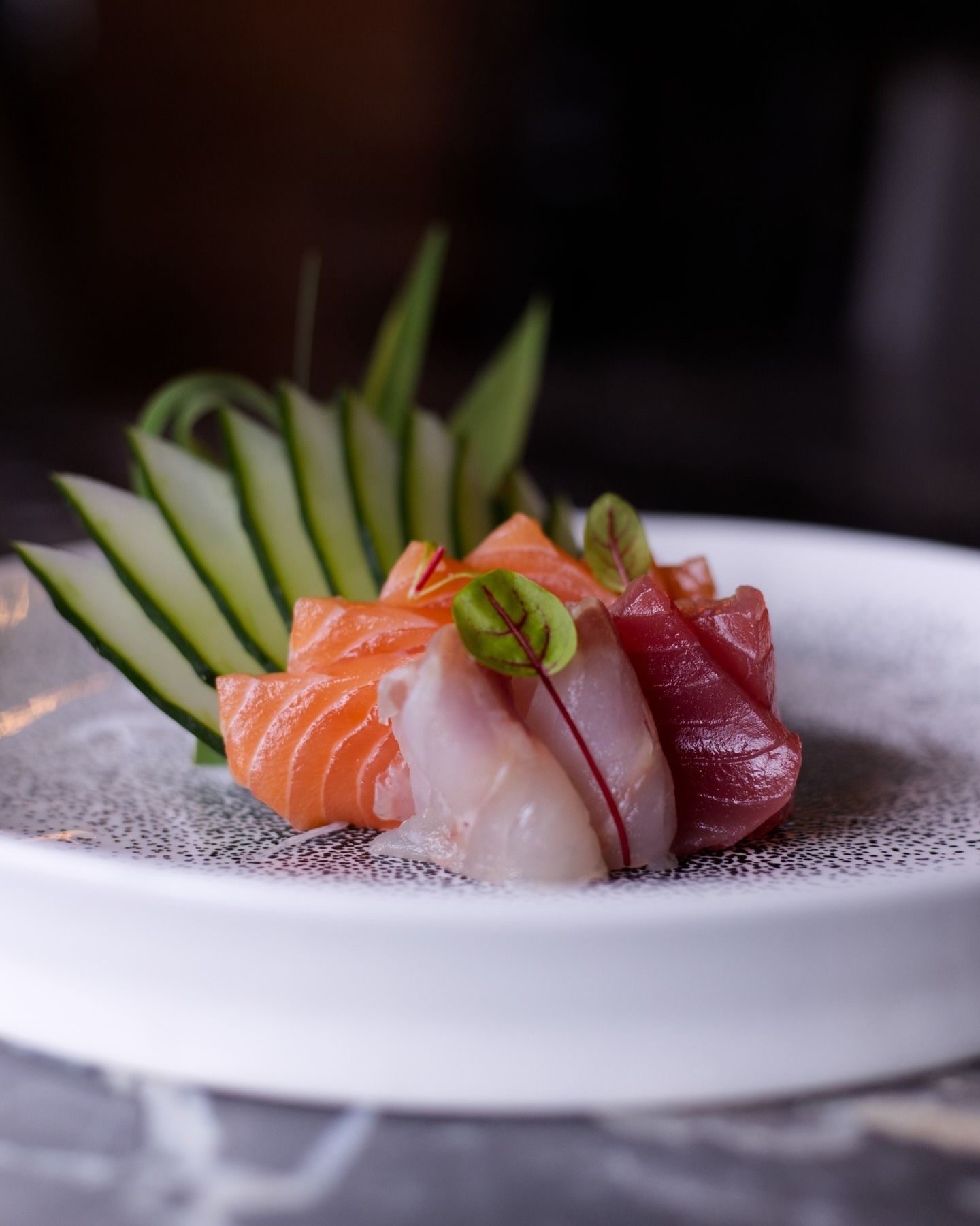 Piatto di sashimi con fette di salmone, pesce bianco e tonno guarnite con cetriolo e un germoglio su un piatto maculato.