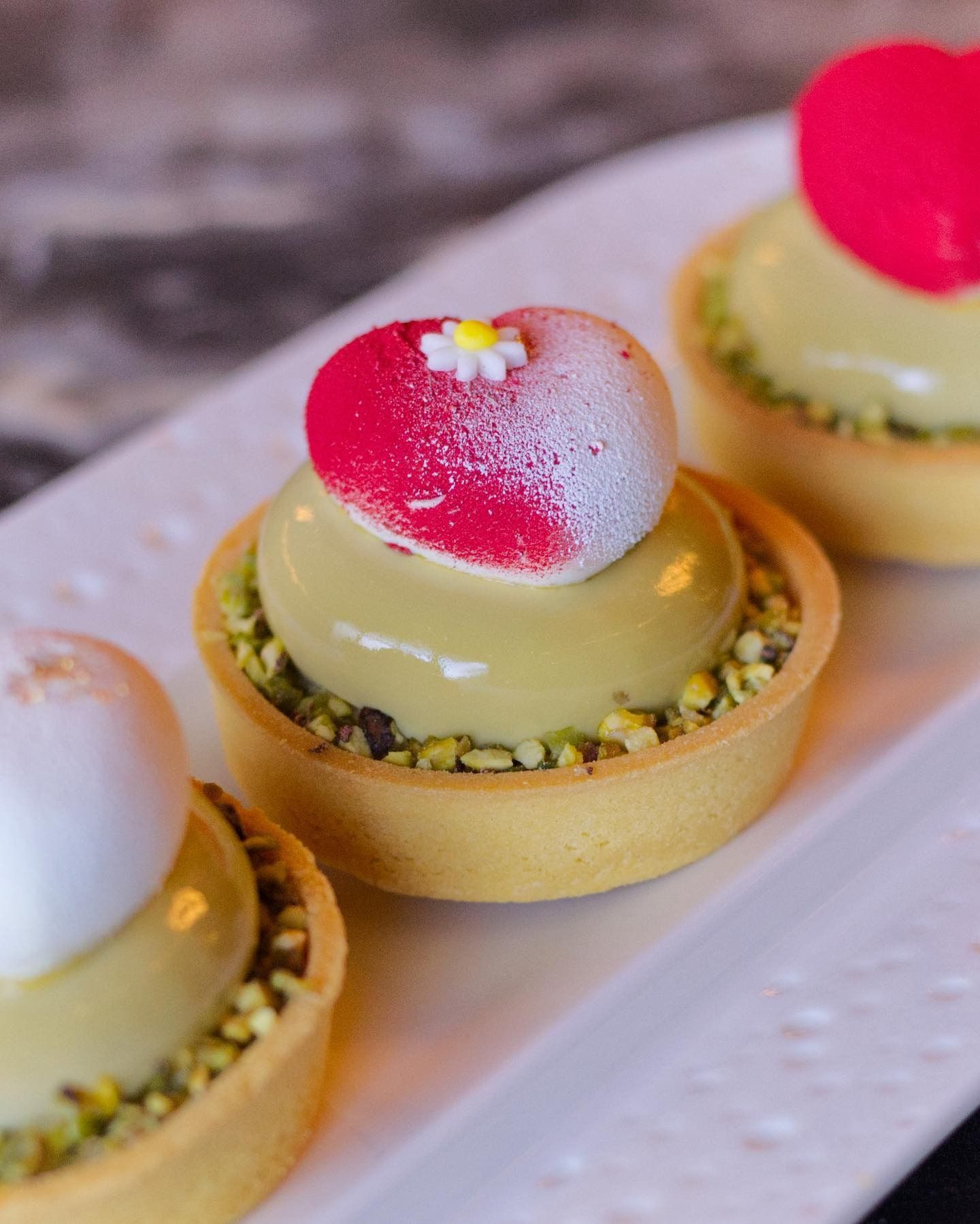 Tre crostatine al pistacchio decorate con mousse rosa a forma di cuore e un minuscolo fiorellino bianco, servite su un piatto bianco.