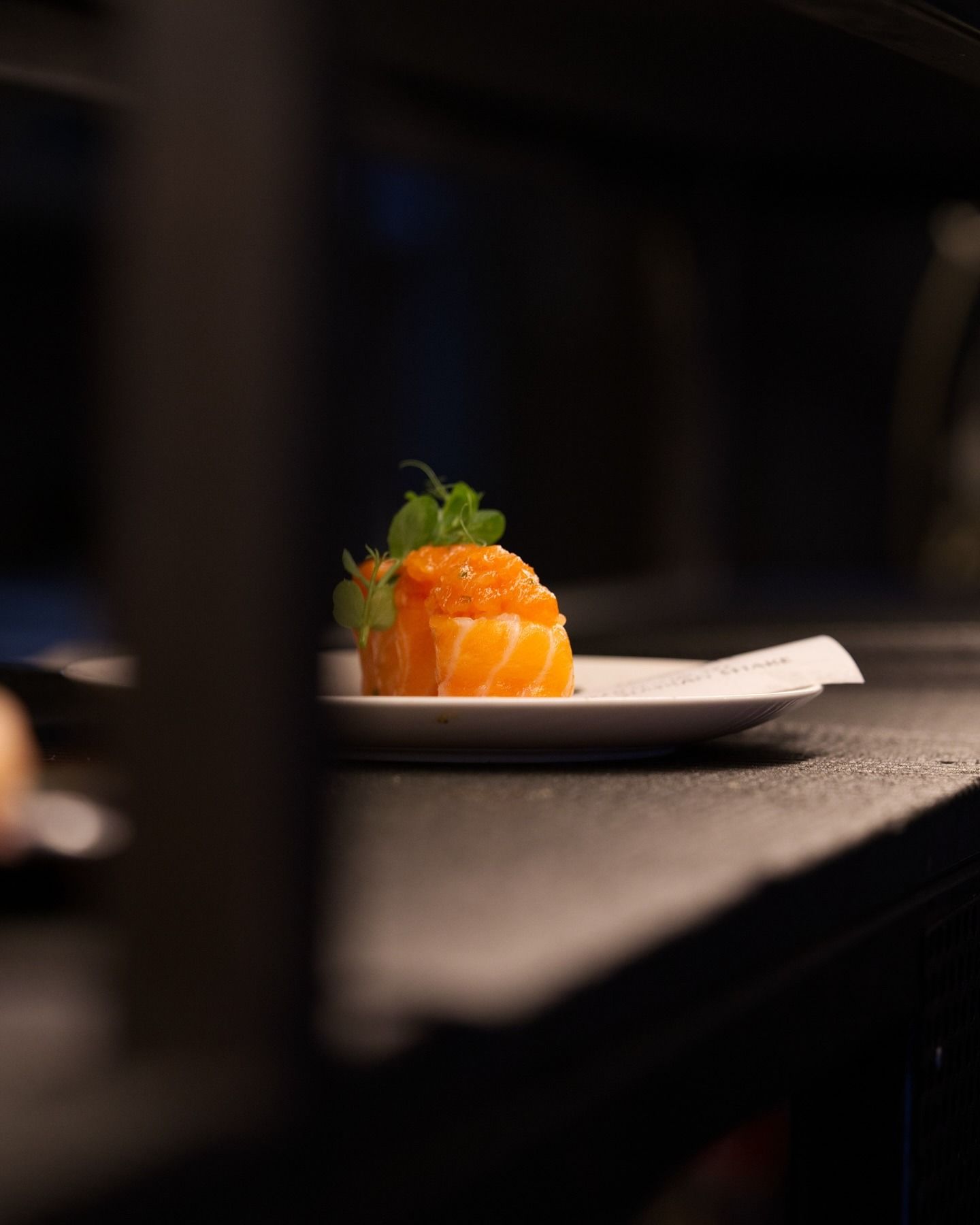 Un singolo pezzo di sushi di salmone guarnito con uova di pesce e un rametto di verdure, servito su un piccolo piatto bianco in un ambiente a luce soffusa.
